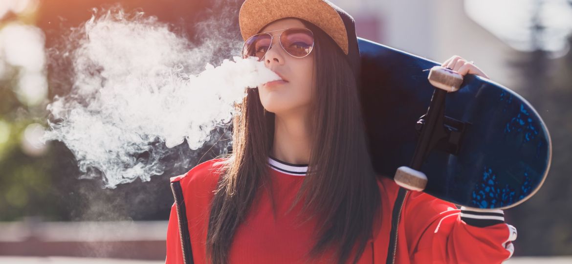 Vaping girl. Young woman with skateboard vape e-cig. Pretty young female in black hat, red clothing vape ecig, vaping device at the sunset. Toned image. Hip-hop style.