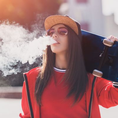 Vaping girl. Young woman with skateboard vape e-cig. Pretty young female in black hat, red clothing vape ecig, vaping device at the sunset. Toned image. Hip-hop style.